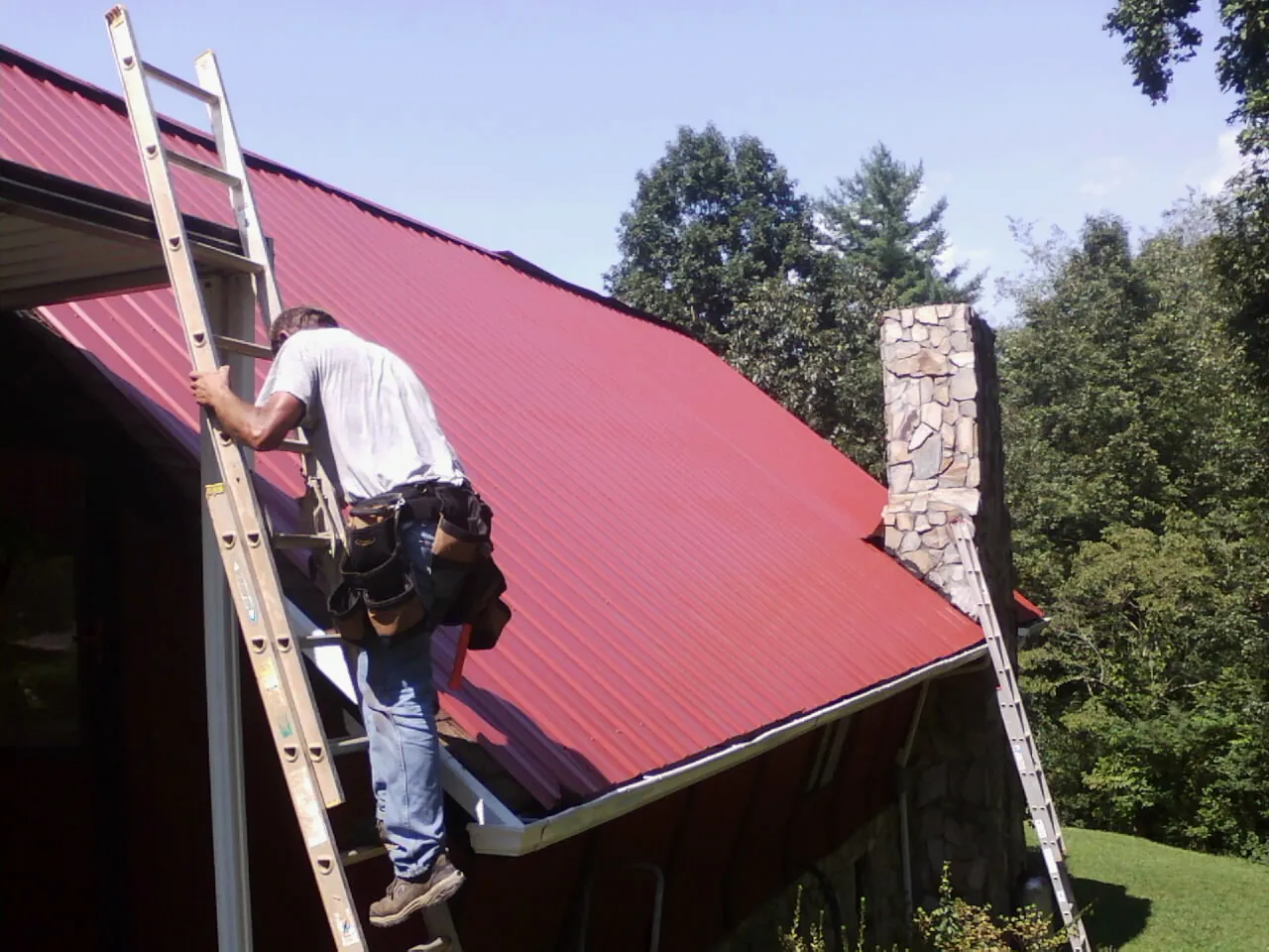 Roofer on a ladder beside a red metal roof and stone chimney — Soffit & Fascia Repair by Roofing Brothers in Rochester Mills